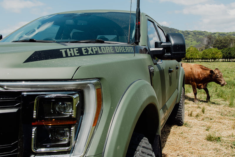 Green pickup truck with 'The Explore Dream' decal on a grassy field with a cow in the background.